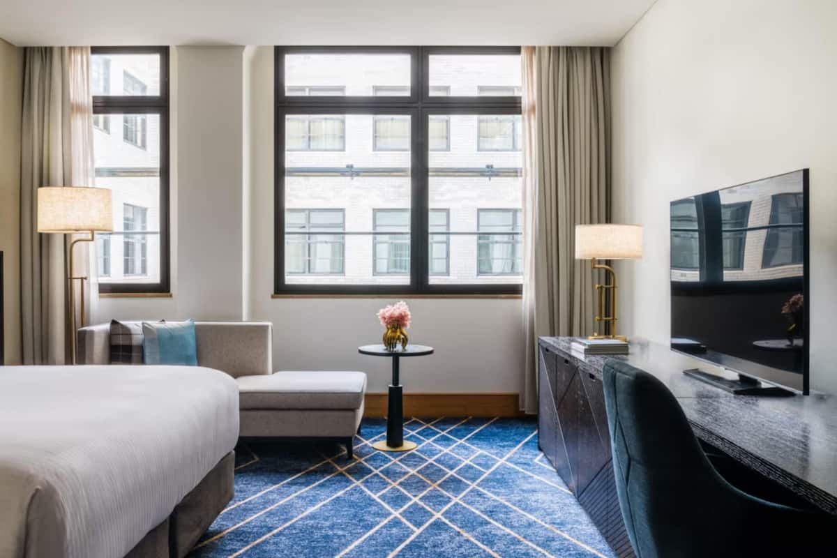 Habitación de hotel con cama, escritorio, silla, pequeña mesa con un florero y televisión.