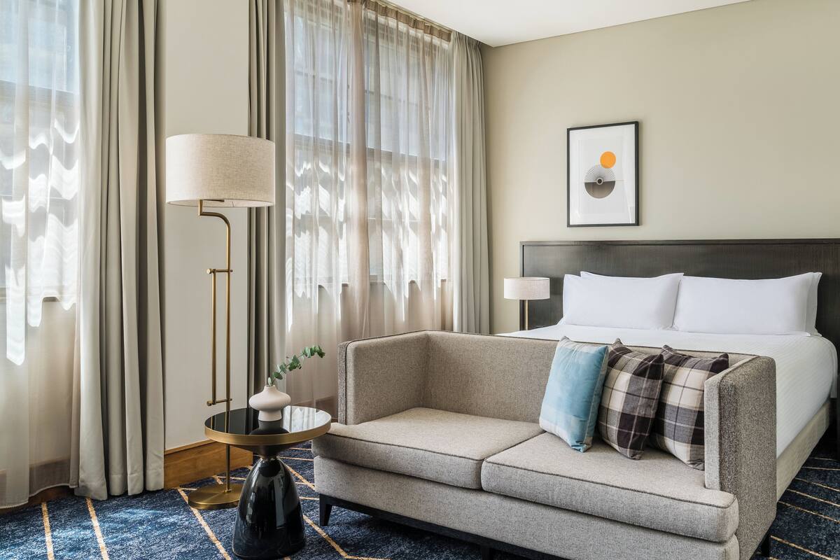 A hotel room with a bed, a sofa, a nightstand, a lamp, and a window with curtains.