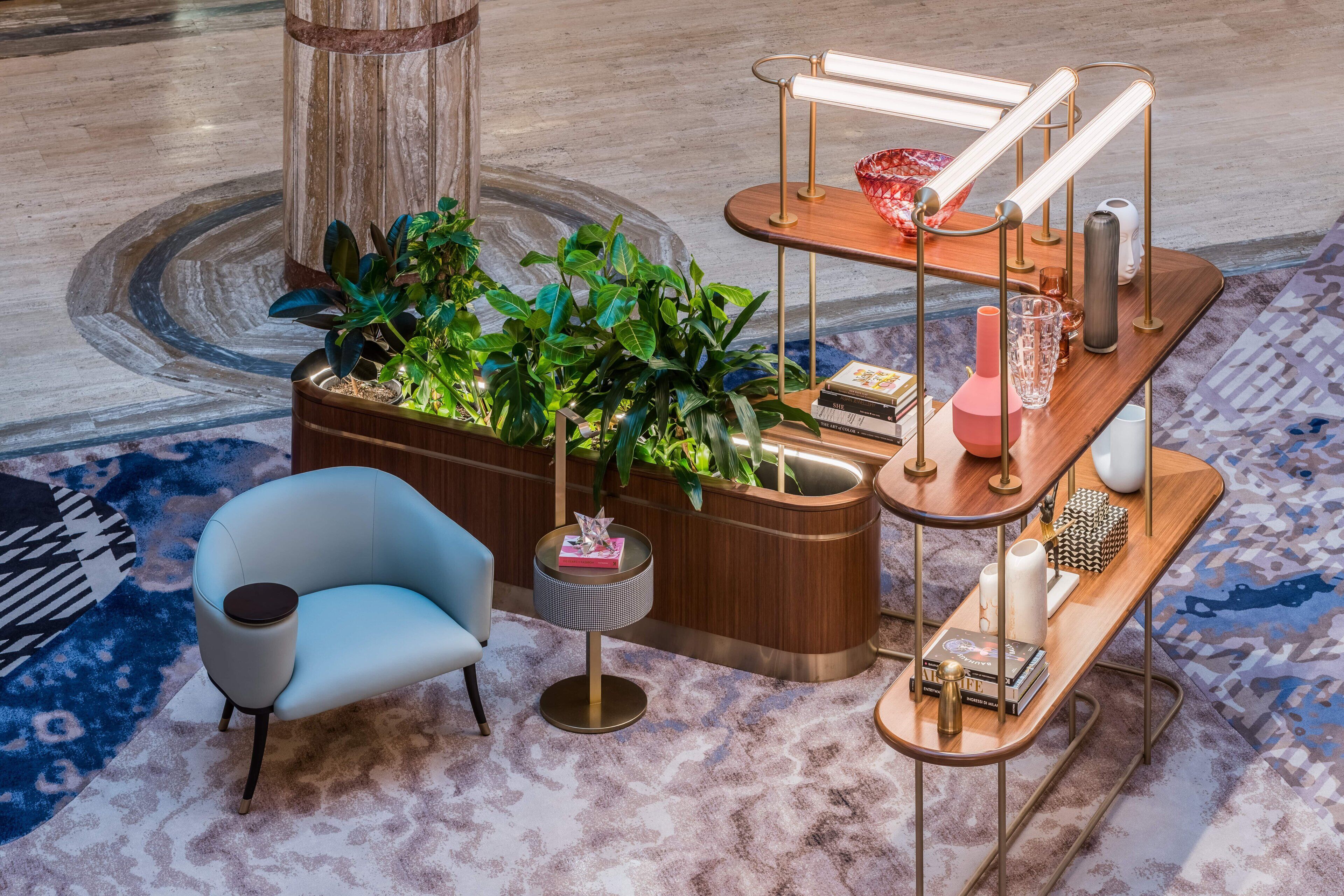 A modern lounge area with a blue armchair, a wooden planter with green plants, a small round table with a lamp, and a wooden side table with decorative items.