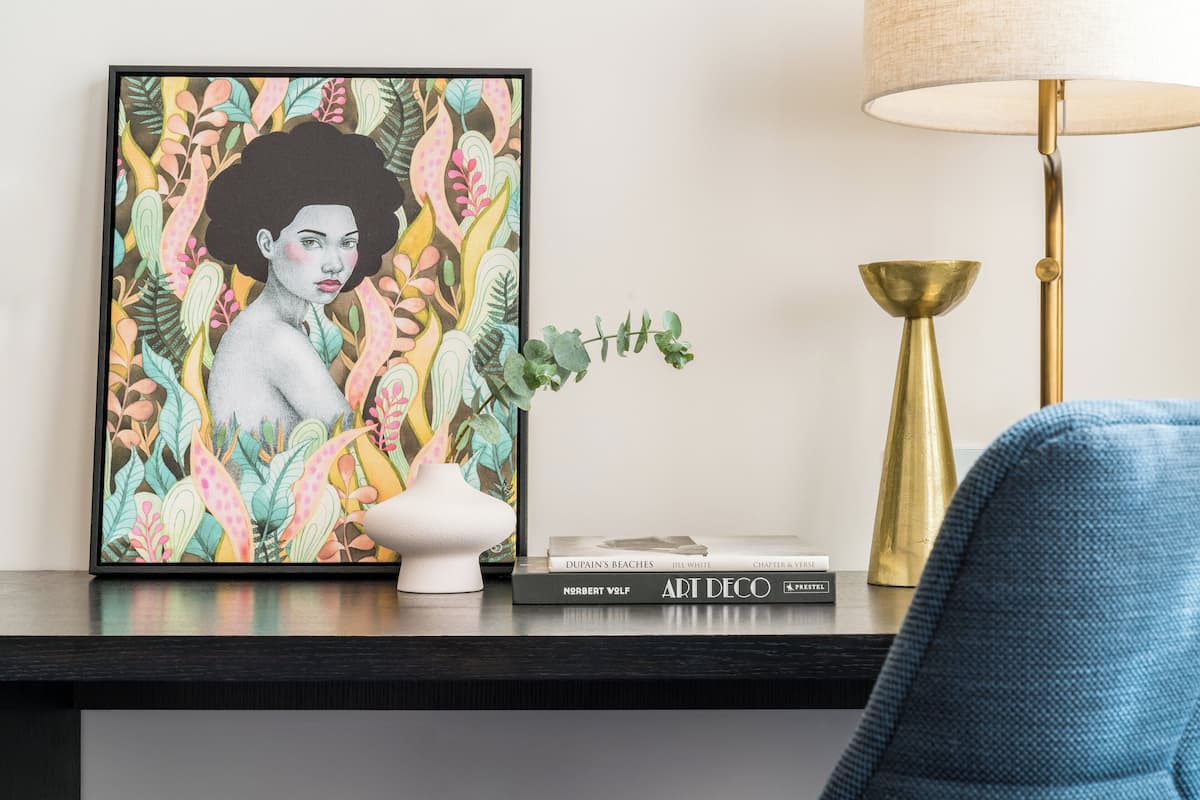 A framed artwork with a floral pattern, a white vase, and a stack of books on a dark wooden surface.