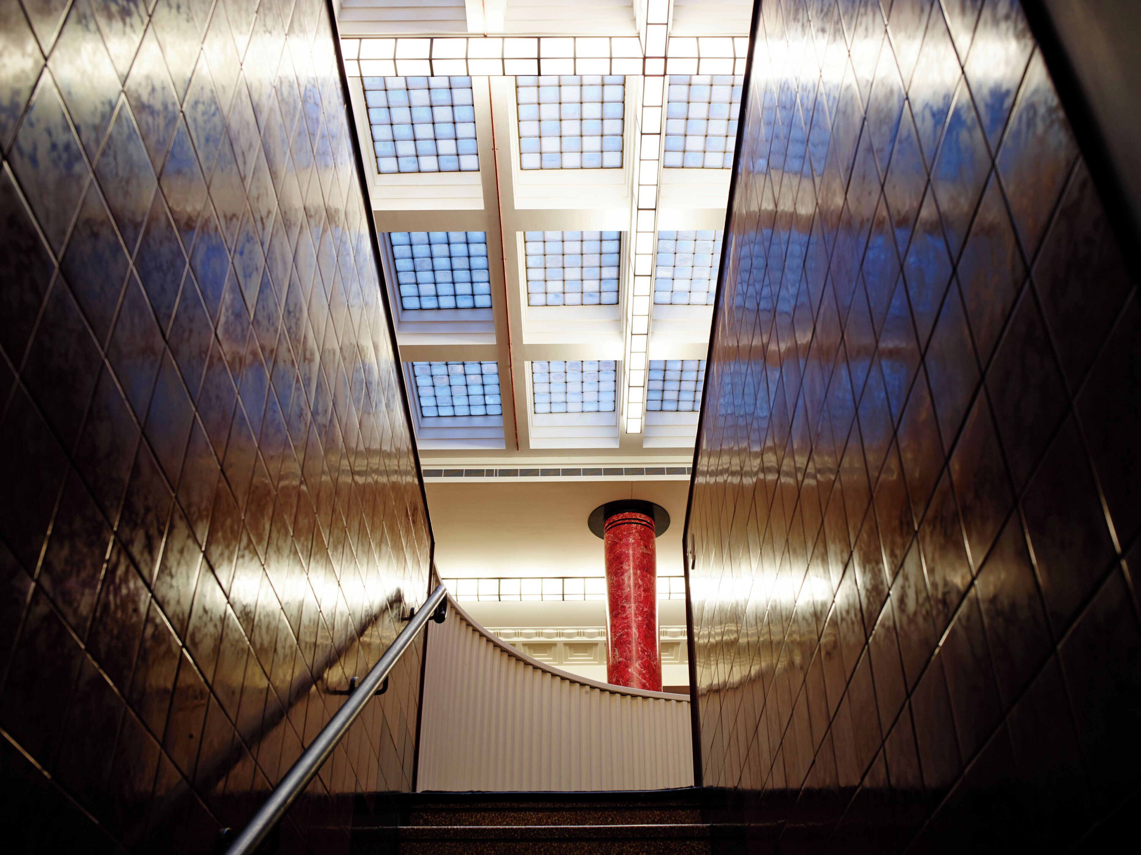 A view of a building's interior with a staircase leading upwards, a red cylindrical column, and a series of windows allowing natural light to enter.