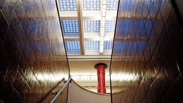 A view of a building's interior with a staircase leading upwards, a red cylindrical column, and a series of windows allowing natural light to enter.