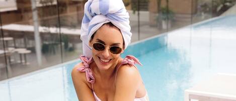 A person sitting by a poolside lounge chair holding a drink.