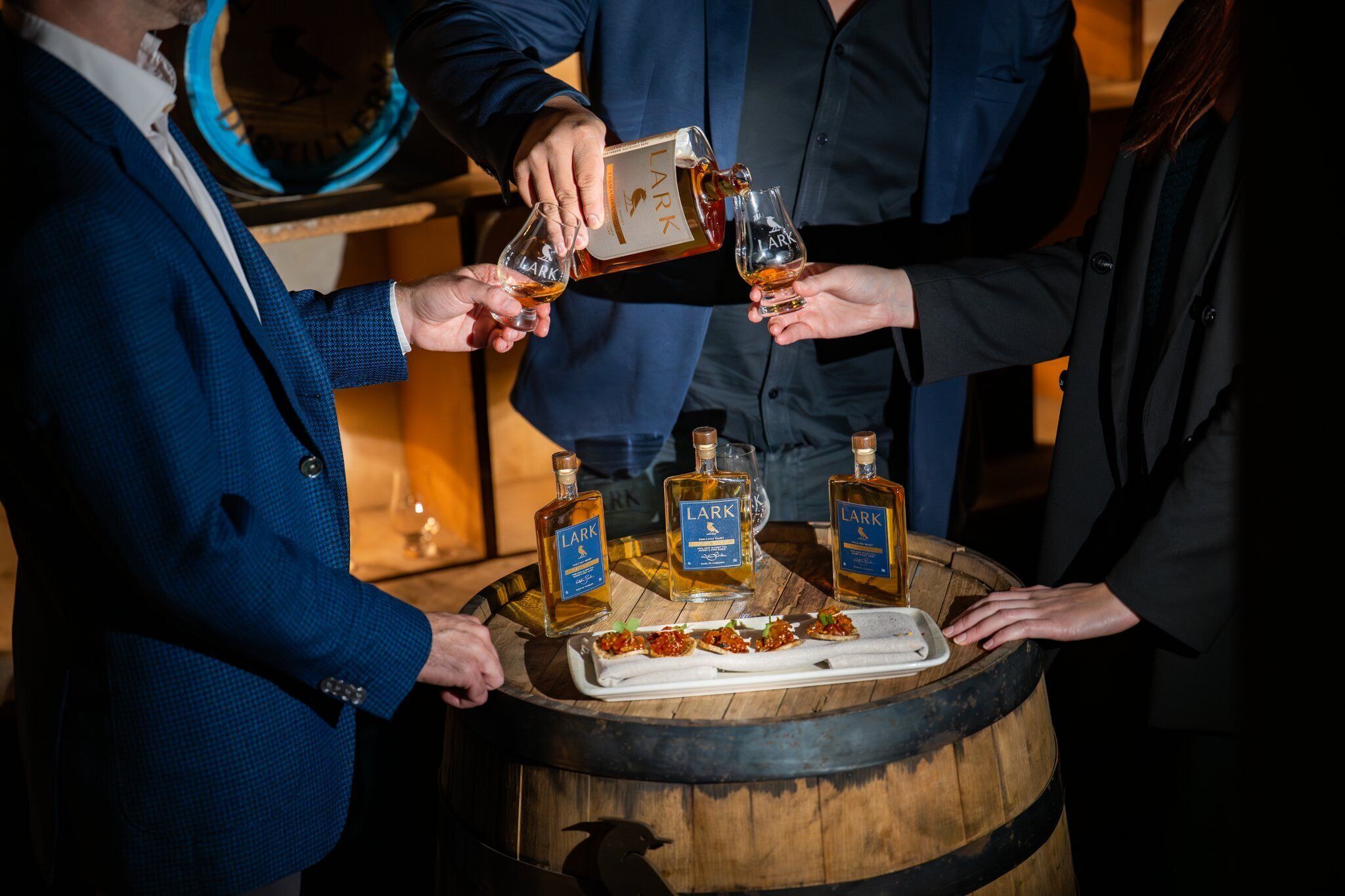 Un groupe de personnes trinquant avec des boissons dans un endroit à la lumière tamisée, avec des bouteilles de whisky Lark et des amuse-gueules disposés sur un tonneau en bois.