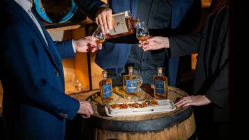 Un groupe de personnes trinquant avec des boissons dans un endroit à la lumière tamisée, avec des bouteilles de whisky Lark et des amuse-gueules disposés sur un tonneau en bois.