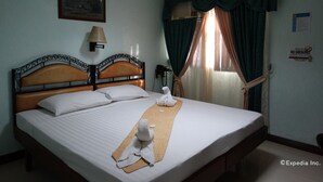 Una cama bien hecha con sábanas blancas y una toalla, una lámpara de pared, un cuadro enmarcado y una ventana con cortinas.
