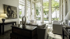 Un salon spacieux avec un plafond haut, de grandes fenêtres et une vue sur l’extérieur. On y trouve un canapé, une table basse et un tableau accroché au mur.