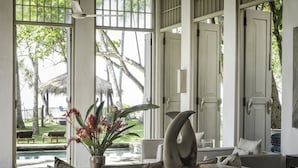 Une pièce avec vue sur une piscine et des palmiers, un canapé avec des coussins, une table avec un vase et un livre, et une grande fenêtre avec des volets blancs.