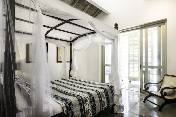A four-poster bed with a patterned bedspread, a chair, and a view of the outside through a glass door. - SATIN DOLL (Unawatuna)