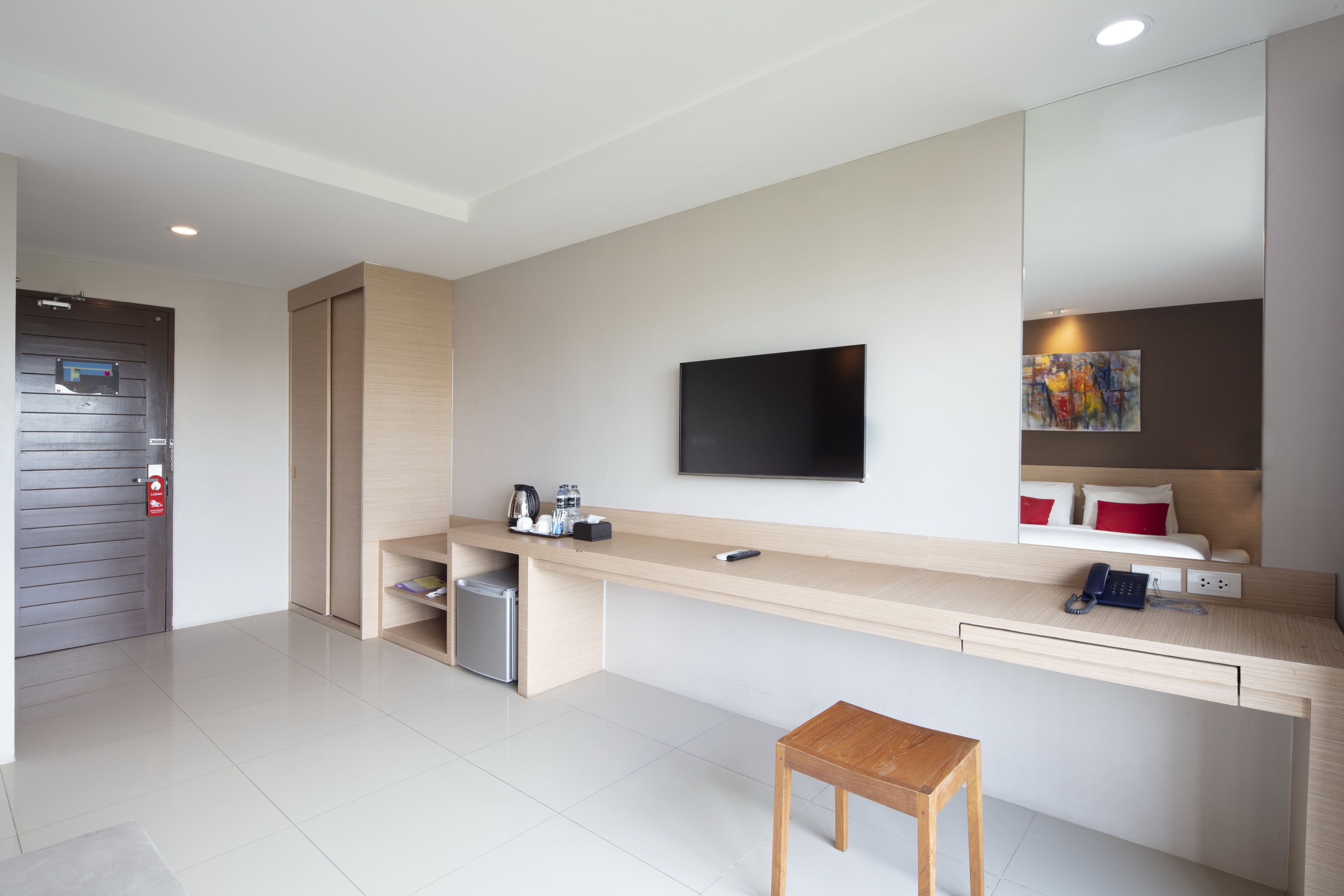 A modern hotel room with a long desk, a small stool, a television mounted on the wall, a mini-fridge, and a painting above the bed.
