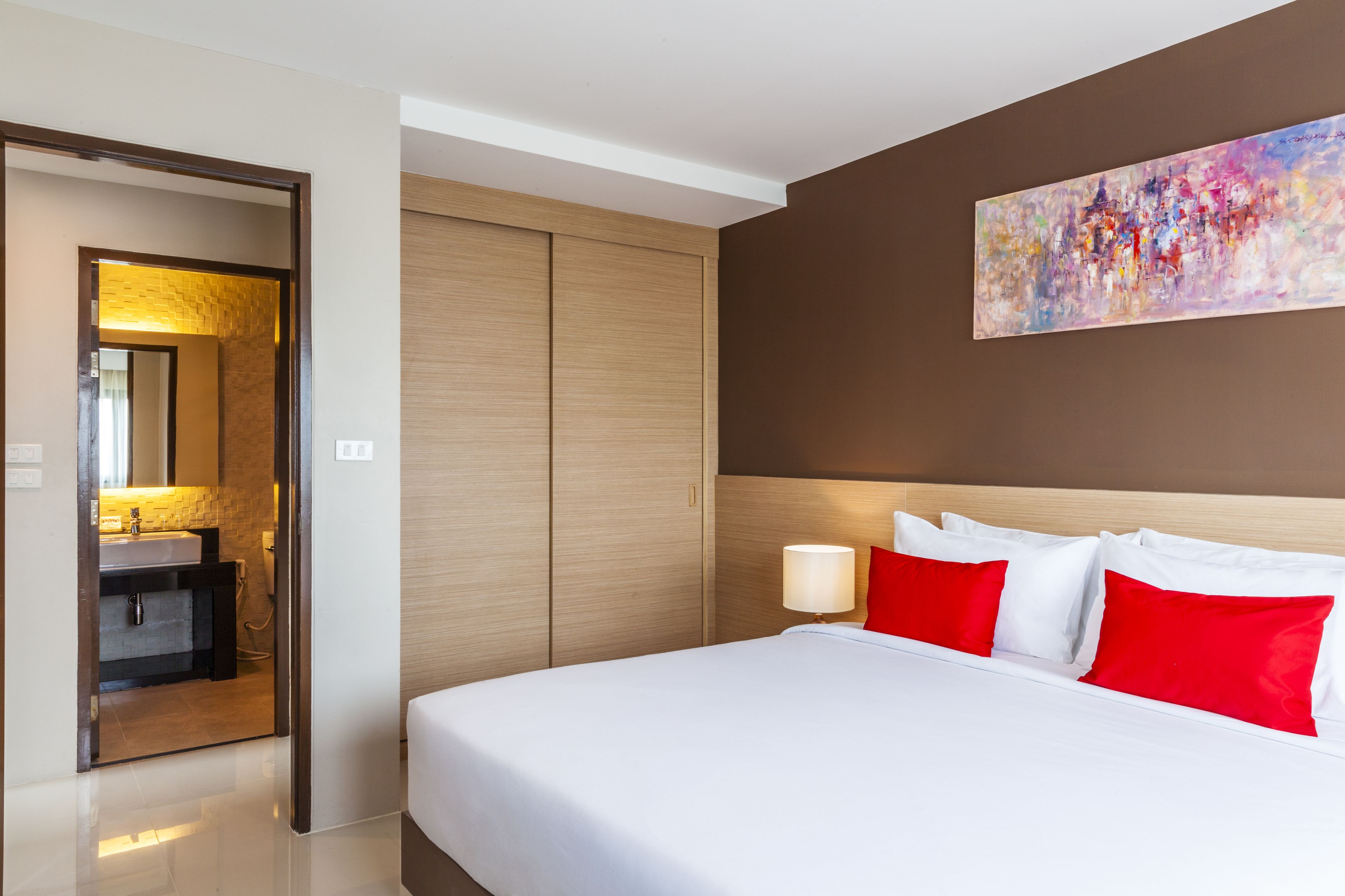Una habitación de hotel con una cama grande, una mesita de noche, un cuadro colgado en la pared y un baño visible a través de una puerta abierta.