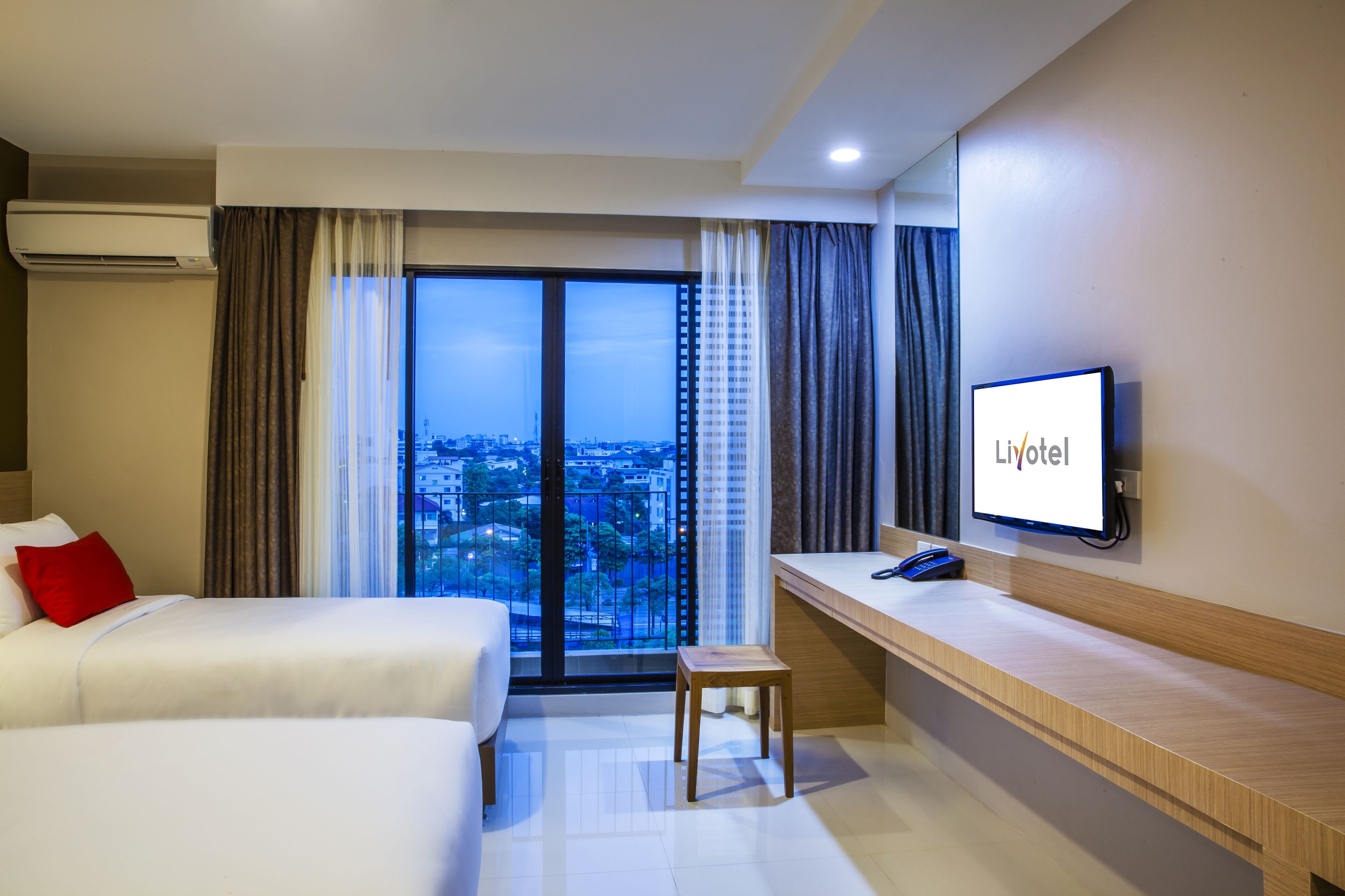 Une chambre d’hôtel avec deux lits, un bureau, un téléviseur affichant le logo « Liyotel » et une vue sur la ville par la fenêtre.