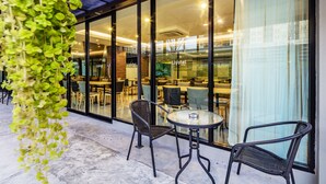 An outdoor seating area with wicker chairs and a glass table, overlooking a restaurant inside a hotel with glass walls. - Livotel Hotel Kaset Nawamin Bangkok (Bangkok)