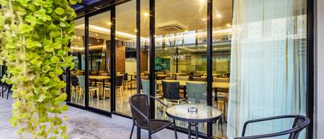 An outdoor seating area with wicker chairs and a glass table, overlooking a restaurant inside a hotel with glass walls.