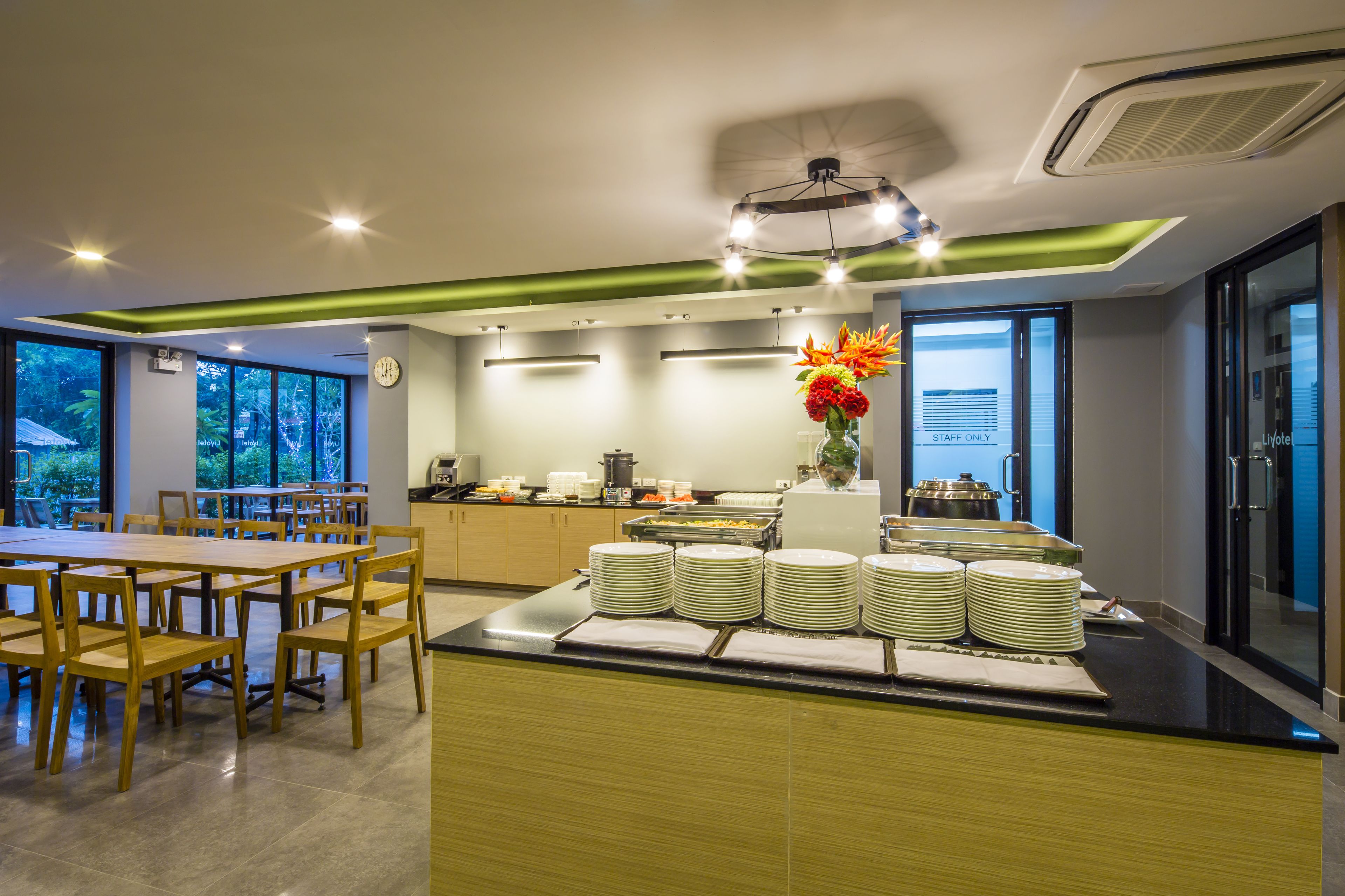A modern dining area with wooden tables and chairs, a buffet counter with stacked plates, and a clock on the wall.
