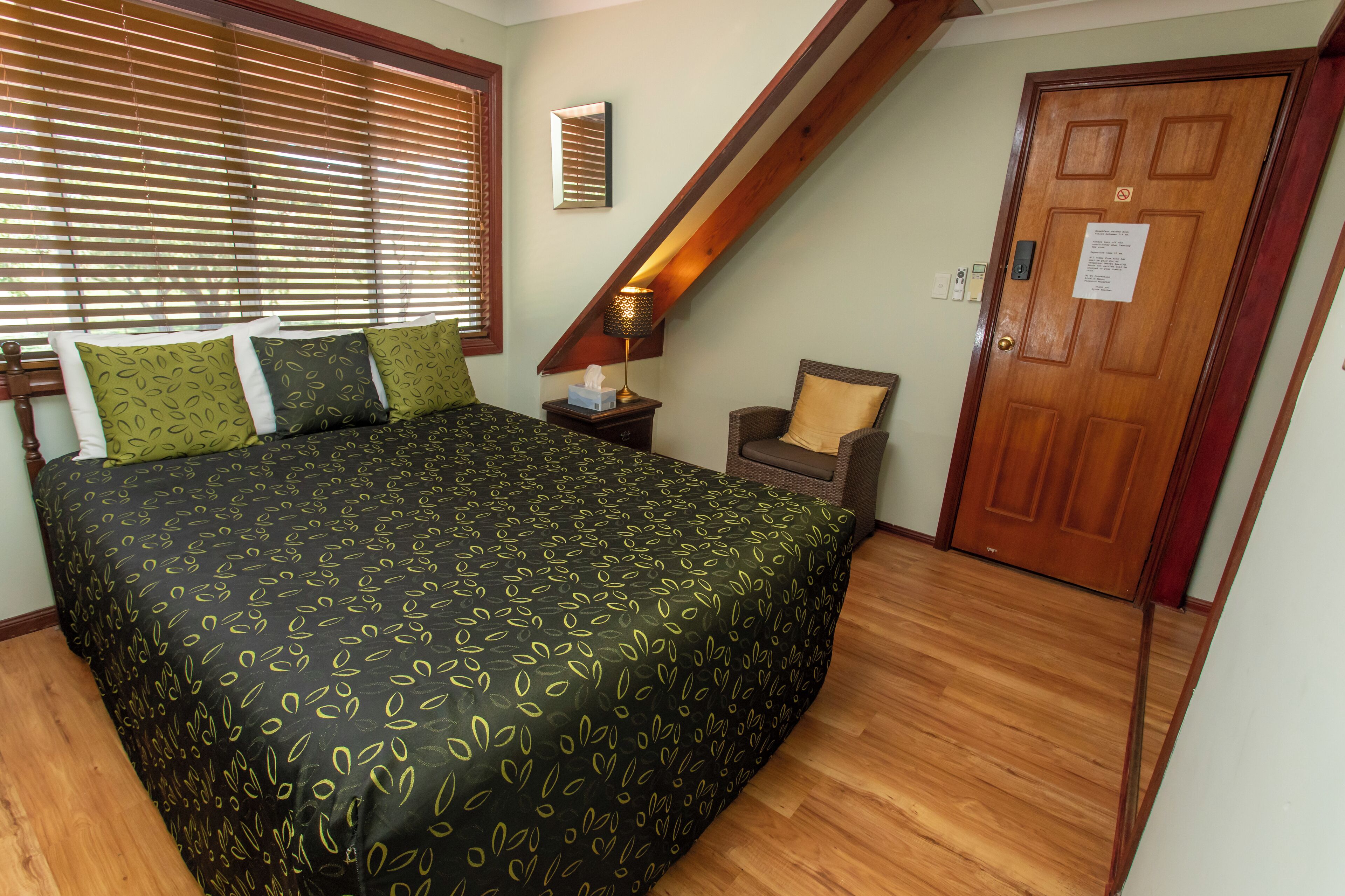 A bedroom with a large bed, a nightstand, a chair, a wooden door, and a window with blinds.