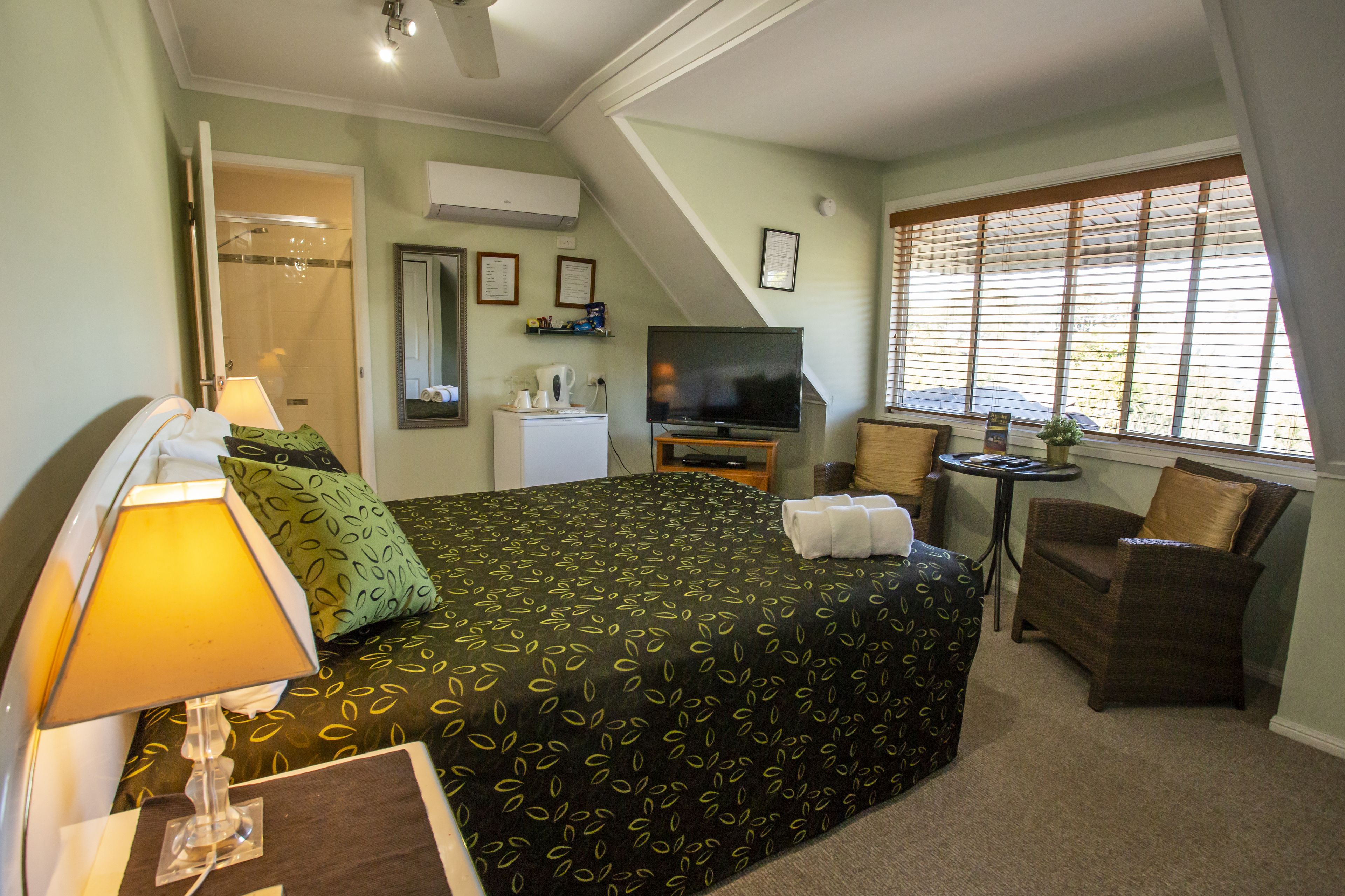 Habitación de hotel con cama, televisión, una mesita, una silla y ventana con persianas.