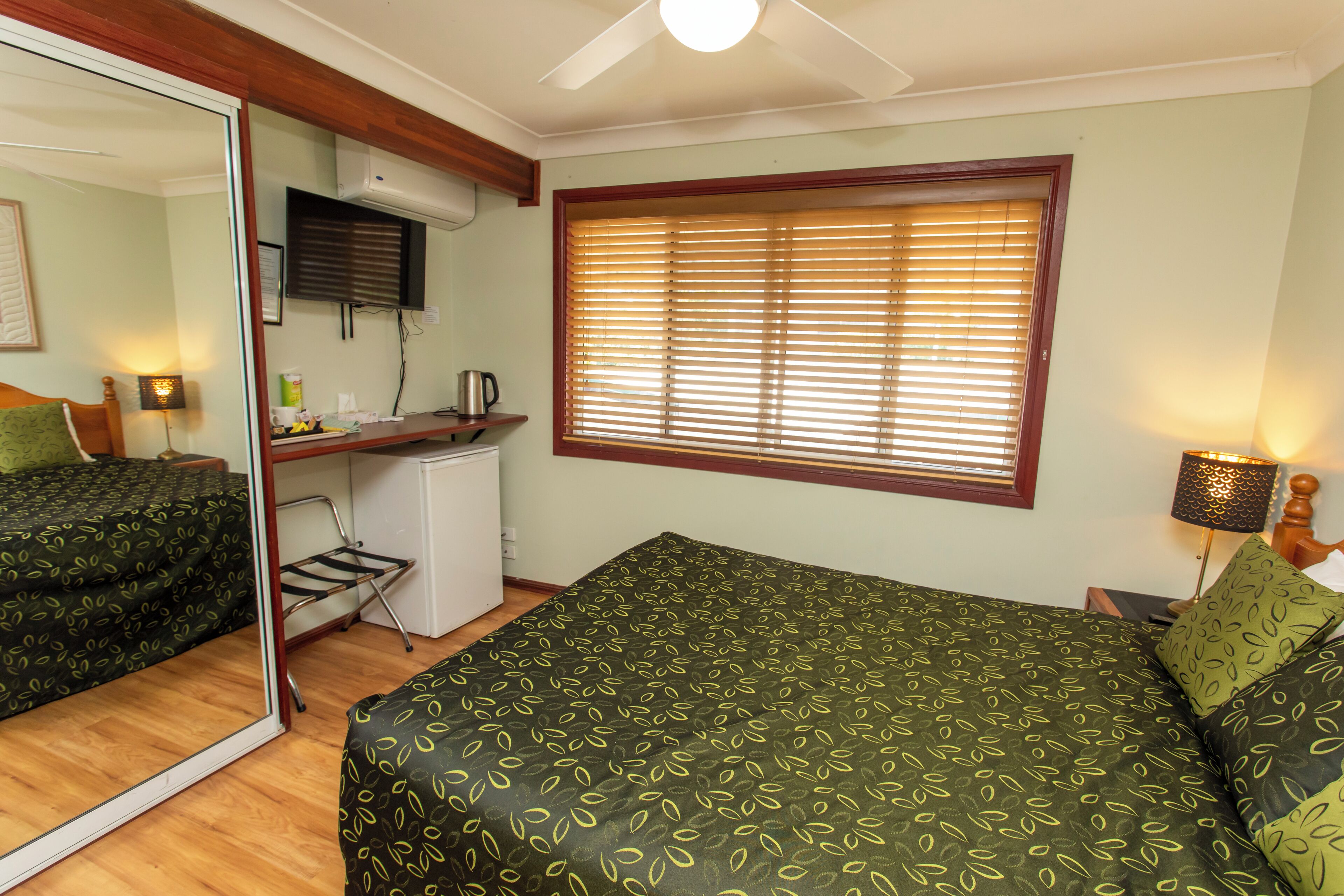 A bedroom with a bed, a desk, a mirror, and a window with blinds.