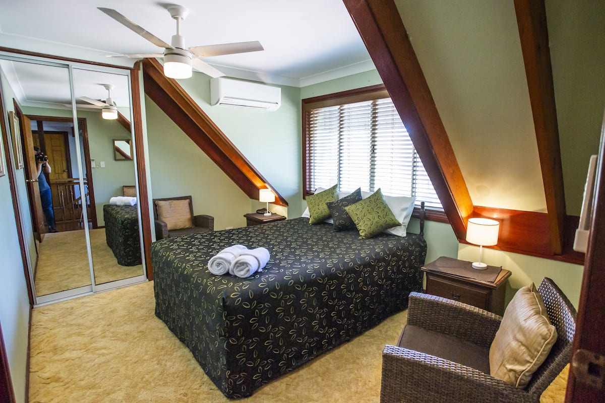 Een slaapkamer met een groot bed, een plafondventilator, een raam met jaloezieën, een nachtkastje met een lamp en een stoel.