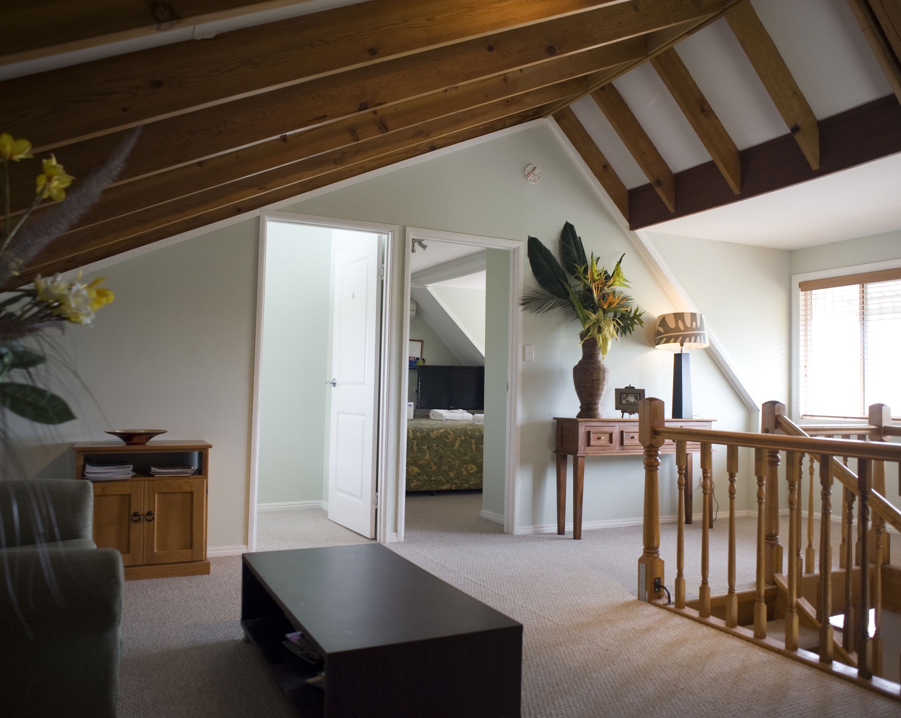 A loft bedroom with a sloped ceiling, a wooden staircase, a bed, a desk, and a potted plant.