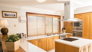 A modern kitchen with wooden cabinets, a built-in microwave, and a central island with a stovetop. - Ainslie Manor Guests House (Redcliffe)