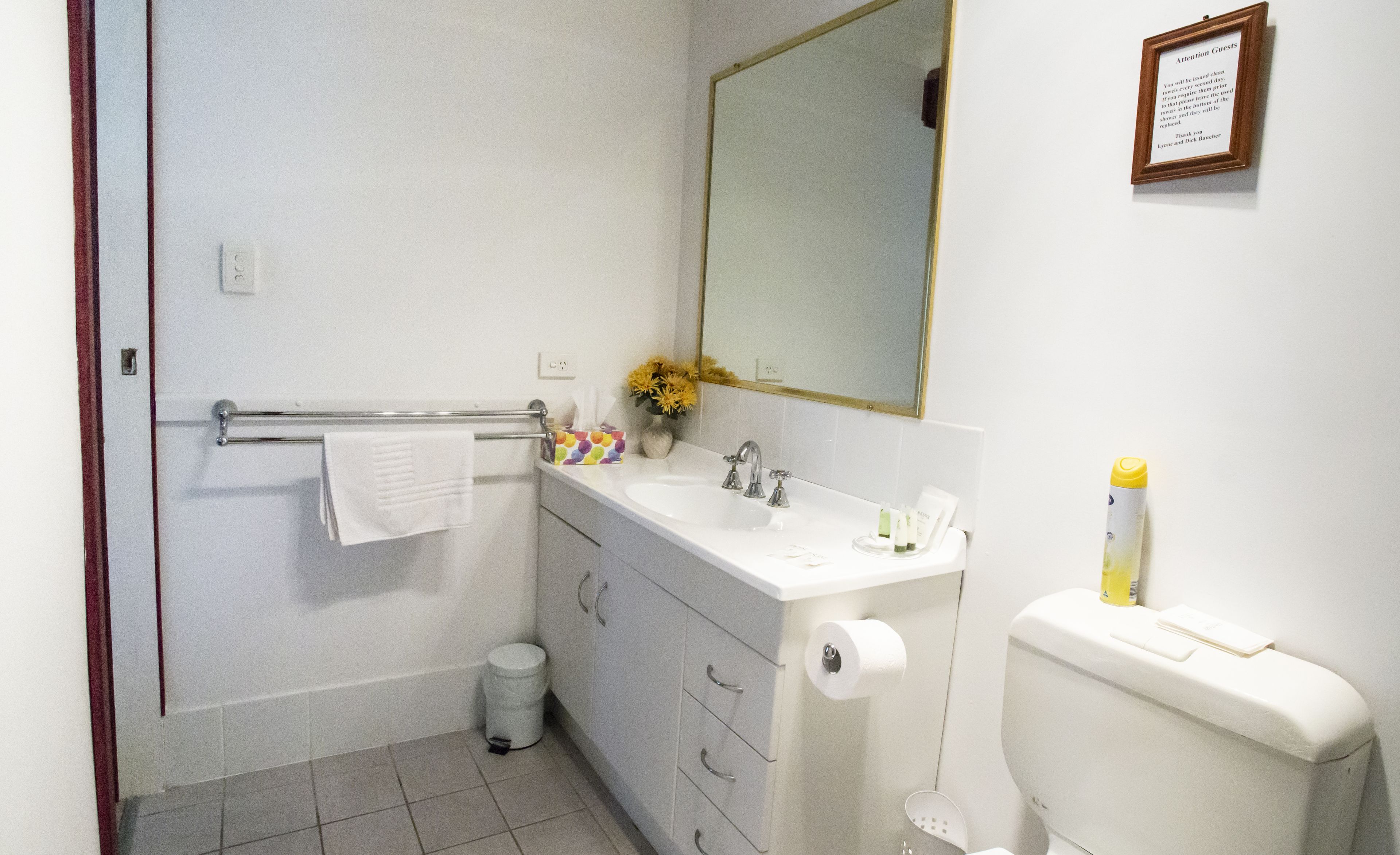 Un baño con lavamanos blanco, espejo e inodoro. Hay un perchero con una toalla blanca, un bote de basura y un peque ño florero con flores amarillas.