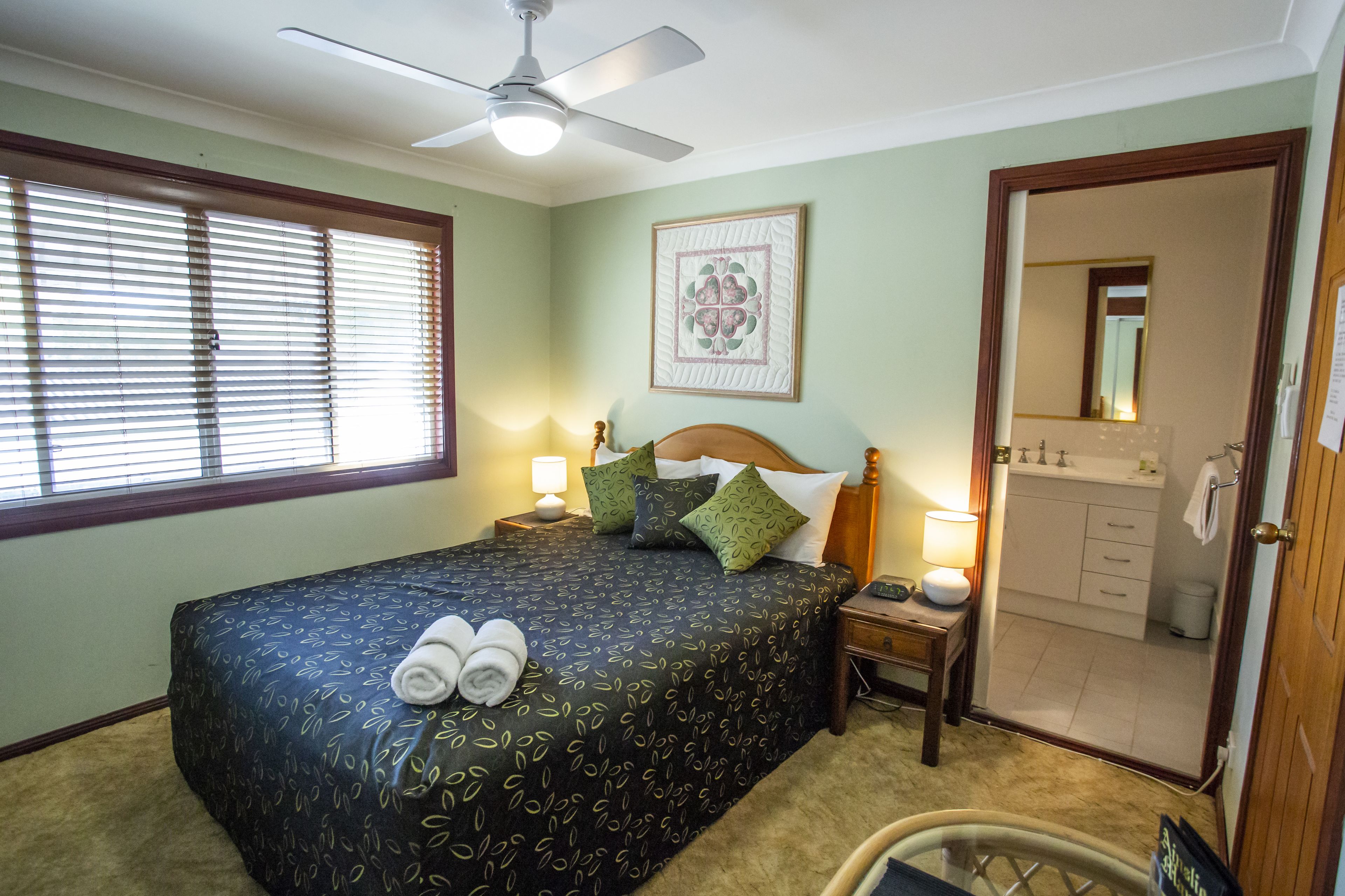 A bedroom with a bed, bedside lamps, a ceiling fan, a window with blinds, a framed artwork, and a bathroom visible through an open door.