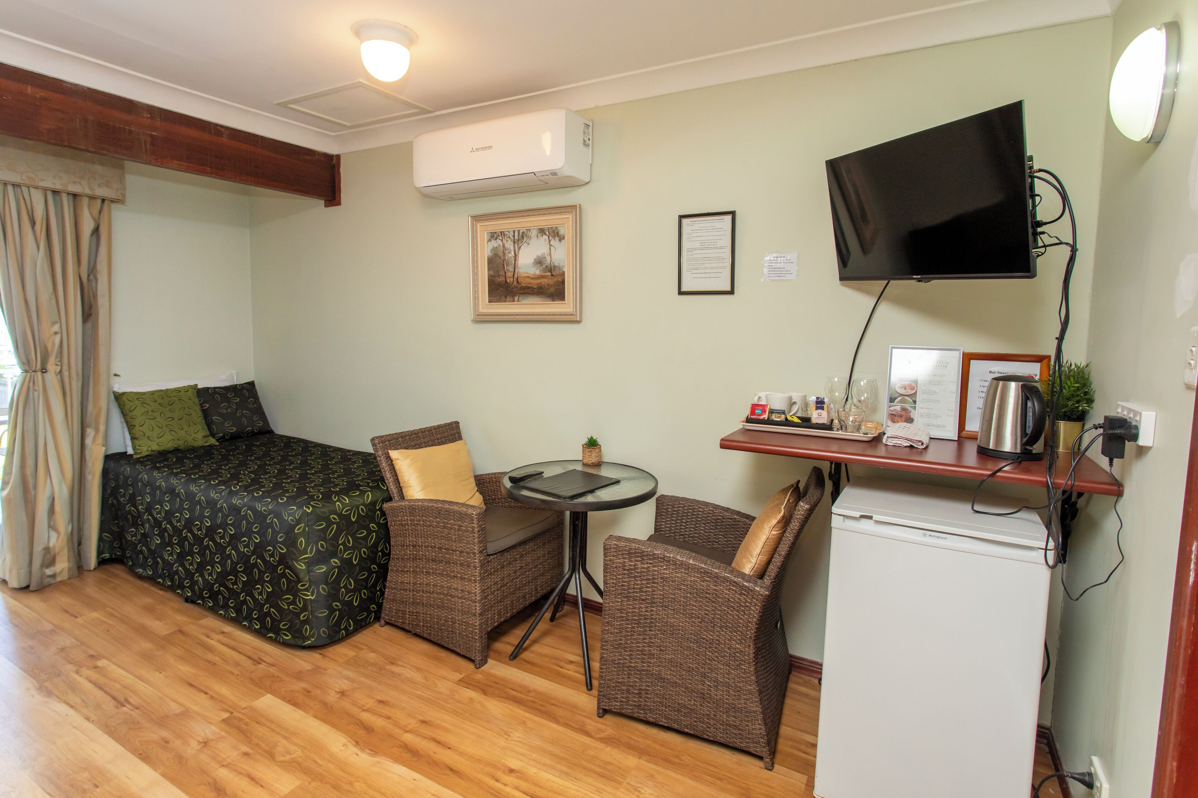 Een kleine kamer met een bed, twee rieten stoelen, een tafeltje, een televisie en een minibar.