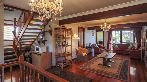 A spacious living room with a wooden staircase, a chandelier, a sofa set, and a coffee table. - Ainslie Manor Guests House (Redcliffe)