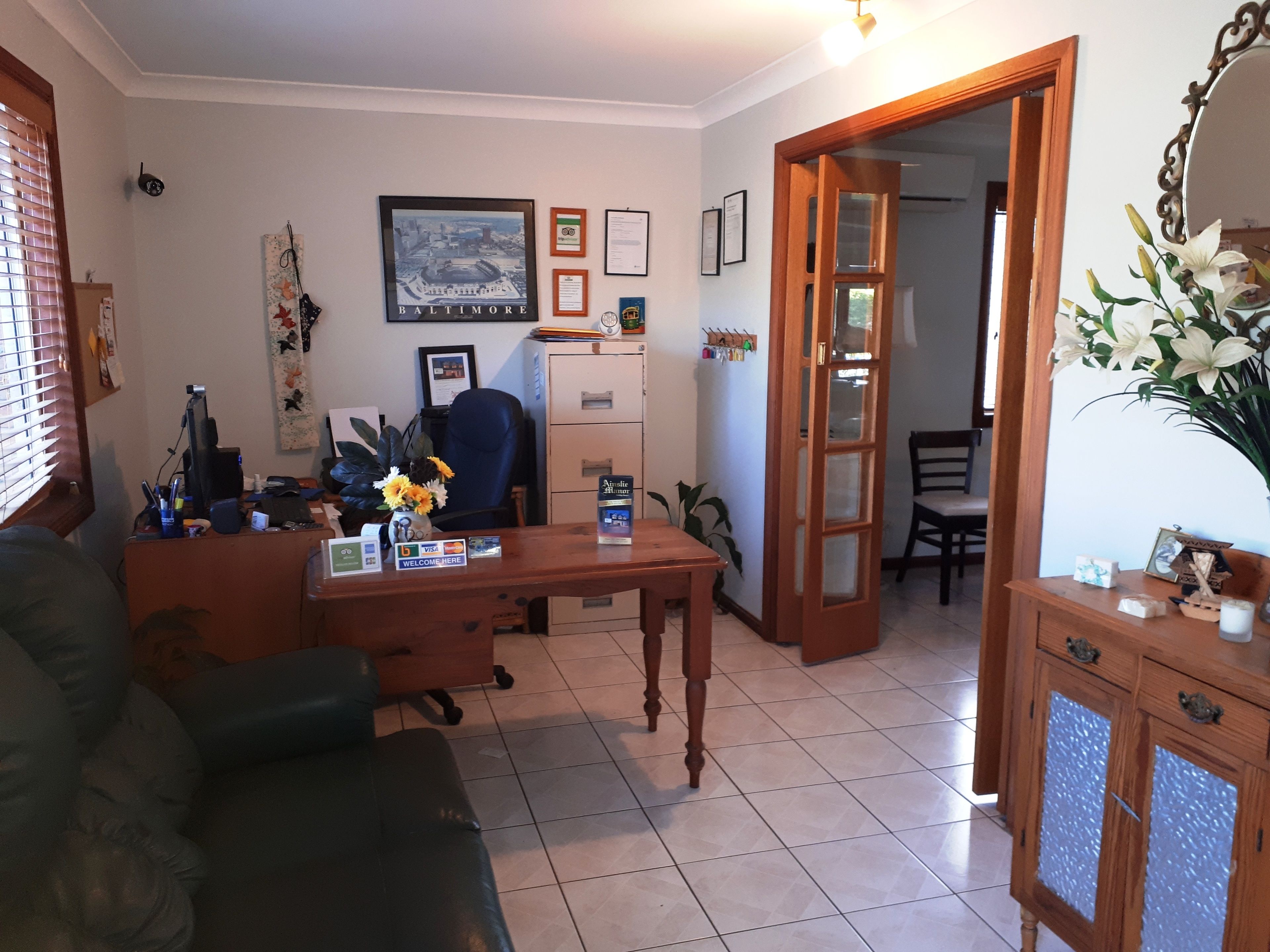 Una oficina en casa con un escritorio, computadora y diversos útiles de oficina. Hay un sofá, una silla y una mesa de comedor con sillas al fondo.