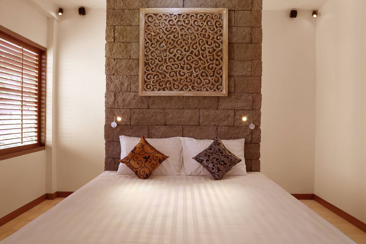 A bedroom with a large bed, two decorative pillows, a stone wall with a carved panel, and a window with blinds.
