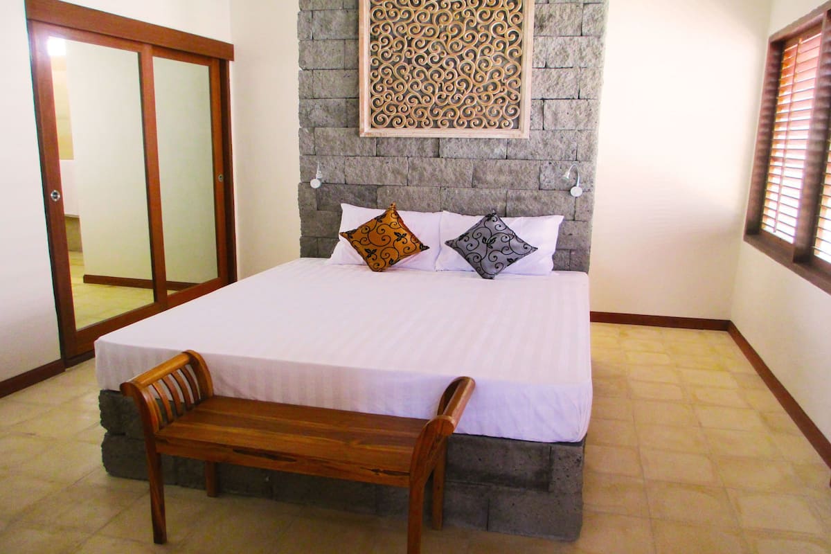 A bedroom with a large bed, a wooden bench, a stone wall, and a patterned wall decoration.