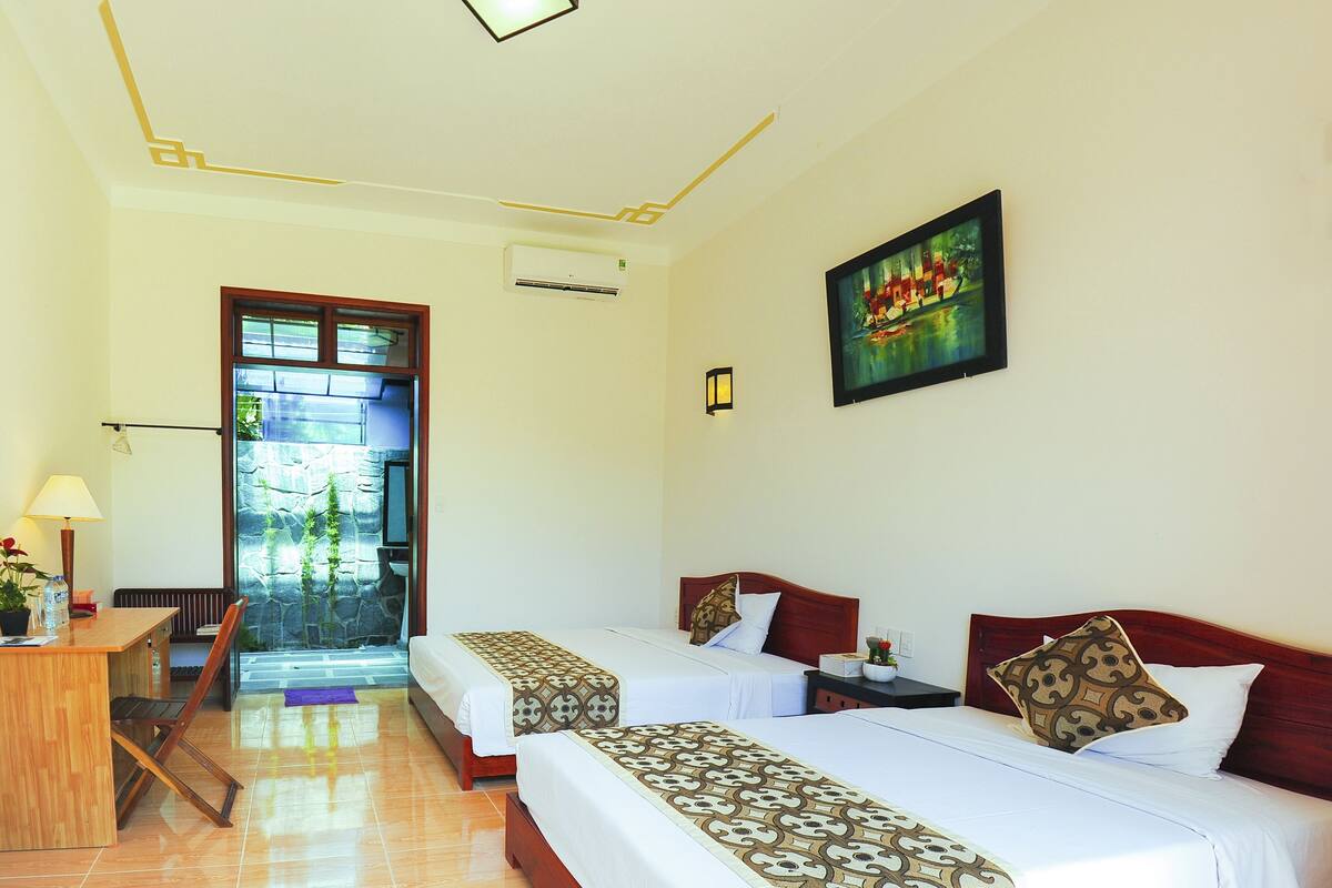 A hotel room with two beds, a desk, a chair, a window with a view of greenery, and a painting on the wall.