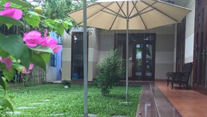 Un espacio al aire libre cubierto con piso de baldosas, un banco y un pequeño jardín con césped y una planta rosada en flor.