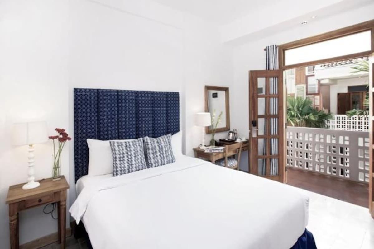 Habitación de hotel con una cama grande, una mesita de noche de madera con una lámpara, un espejo y vistas al exterior a través de una puerta de cristal.