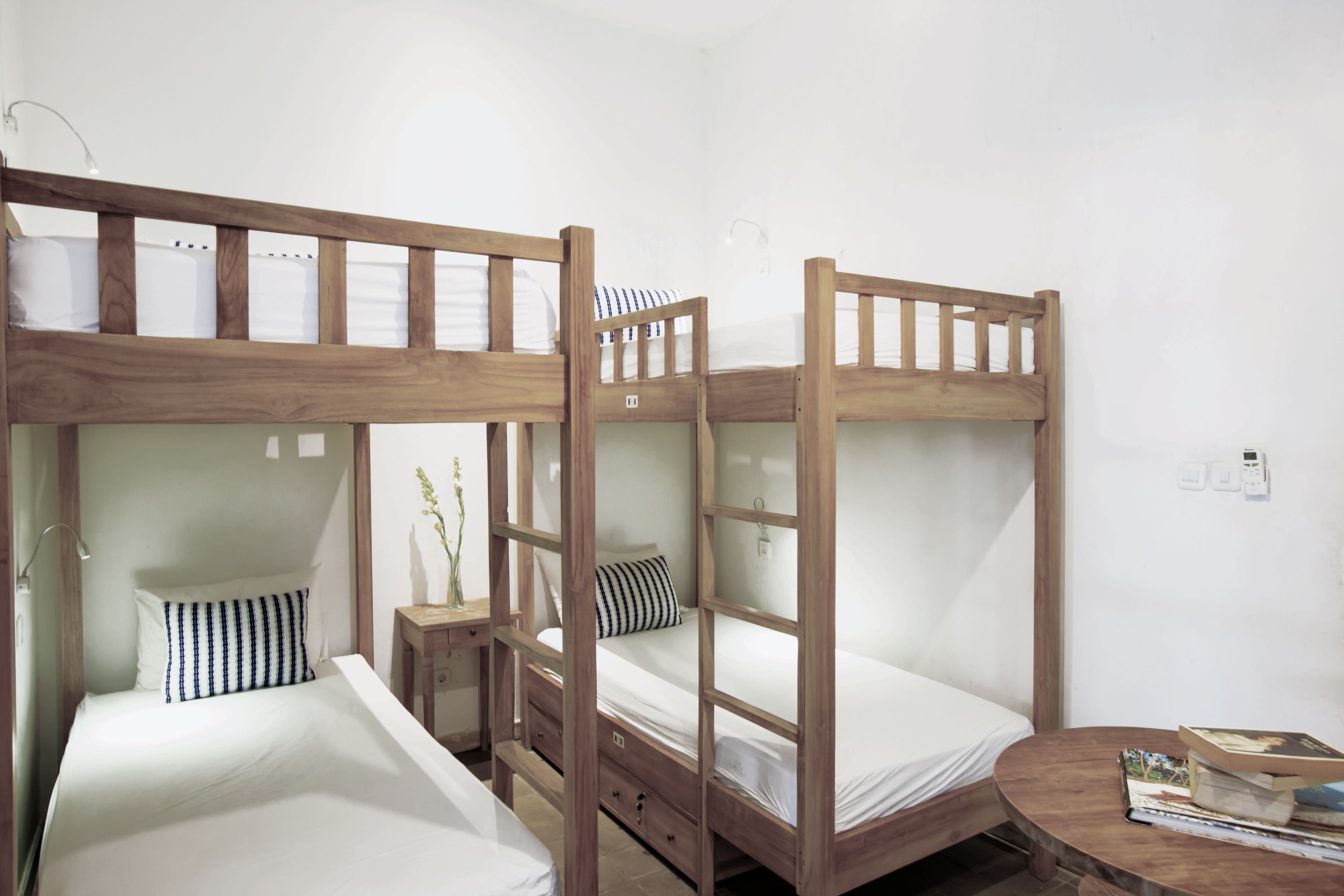 Een slaapkamer met stapelbedden, houten frames, witte beddengoed en een klein nachtkastje met een boek en een vaas.