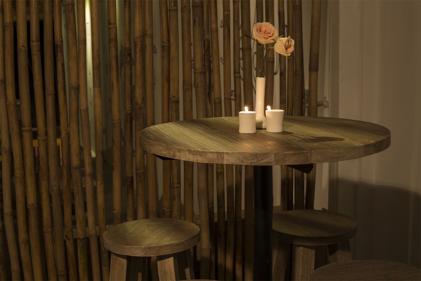 A round wooden table with two candles and a vase with flowers, set against a bamboo backdrop.