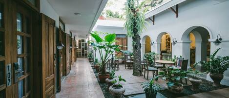 A corridor with wooden doors, numbered 107, leading to a courtyard with potted plants, tables, and chairs.