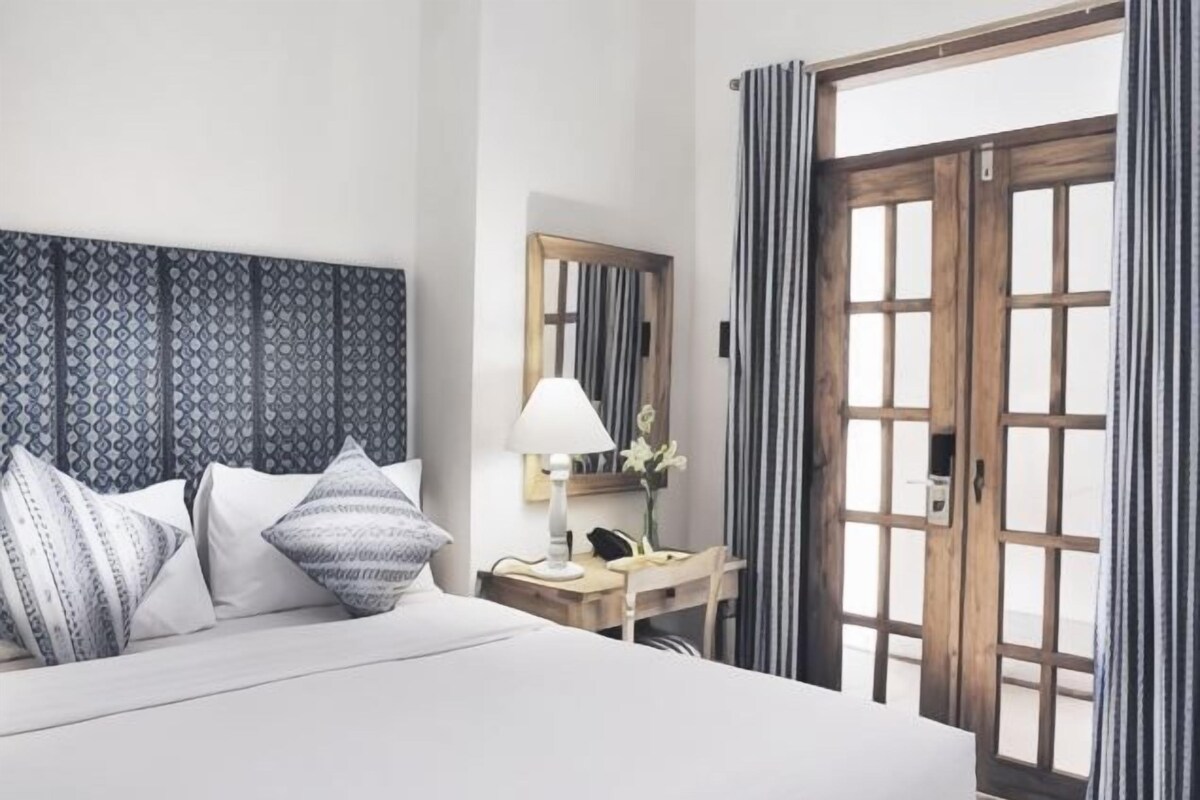 Habitación de hotel con cama, mesita de noche, lámpara y vistas a través de una puerta de madera.