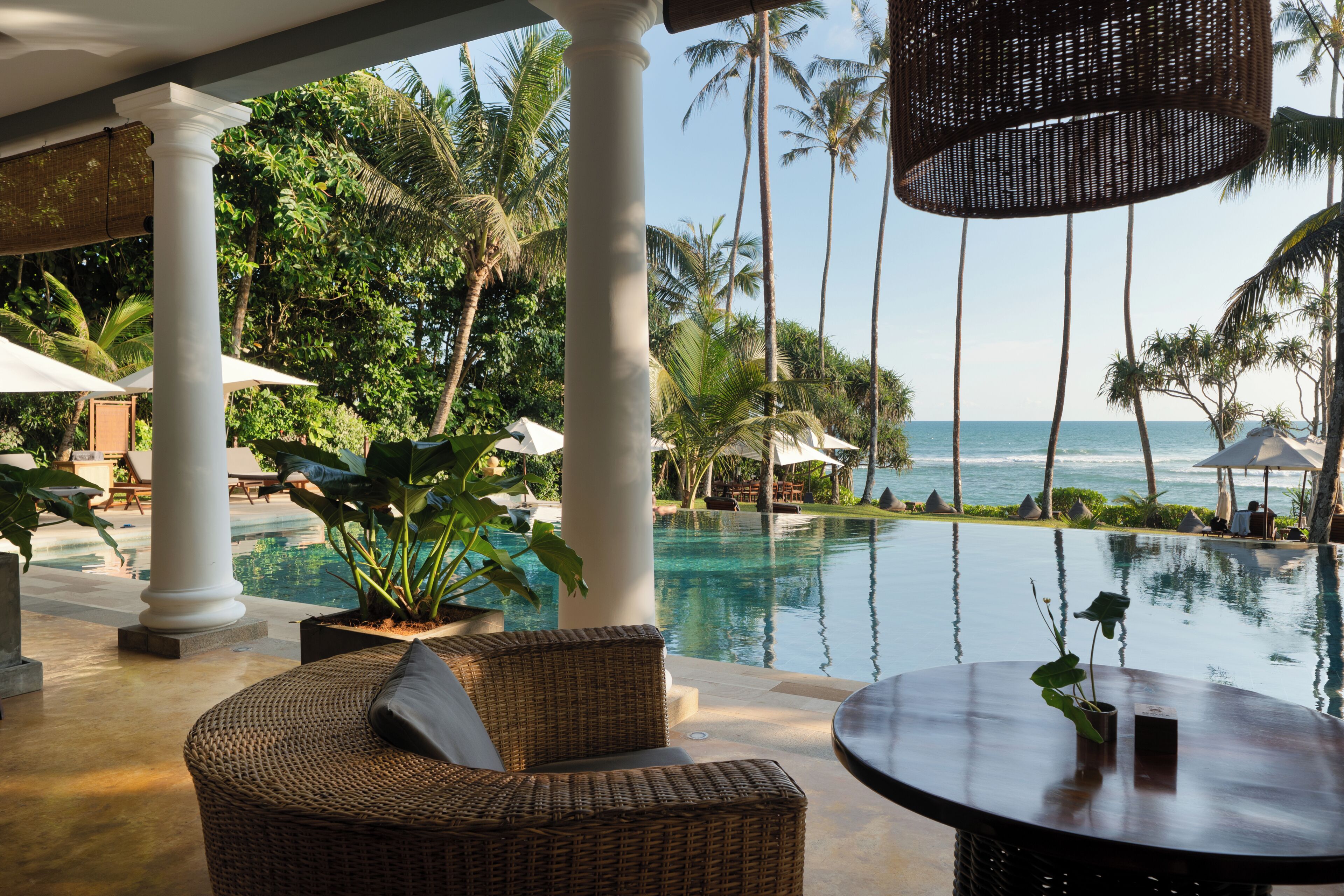 A poolside area with wicker furniture, palm trees, and a view of the ocean.