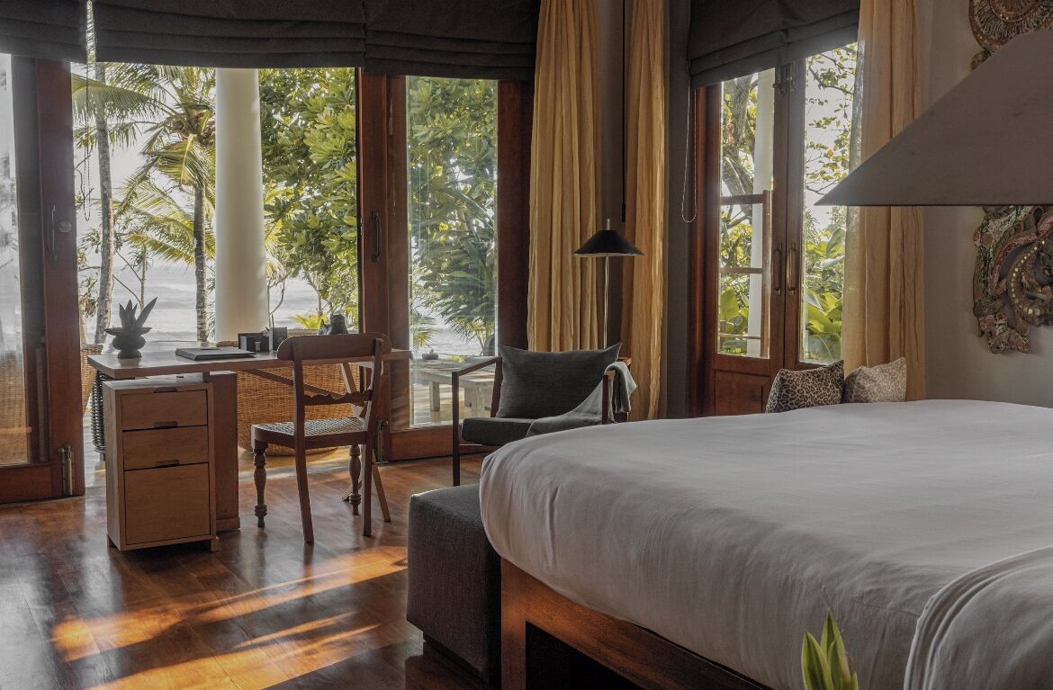 Ein Schlafzimmer mit einem großen Bett, einem Schreibtisch mit Stuhl und Blick auf Palmen durch große Fenster.
