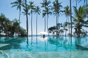 Una piscina azul cristalina rodeada de altas palmeras, con una persona relajándose bajo una sombrilla blanca.