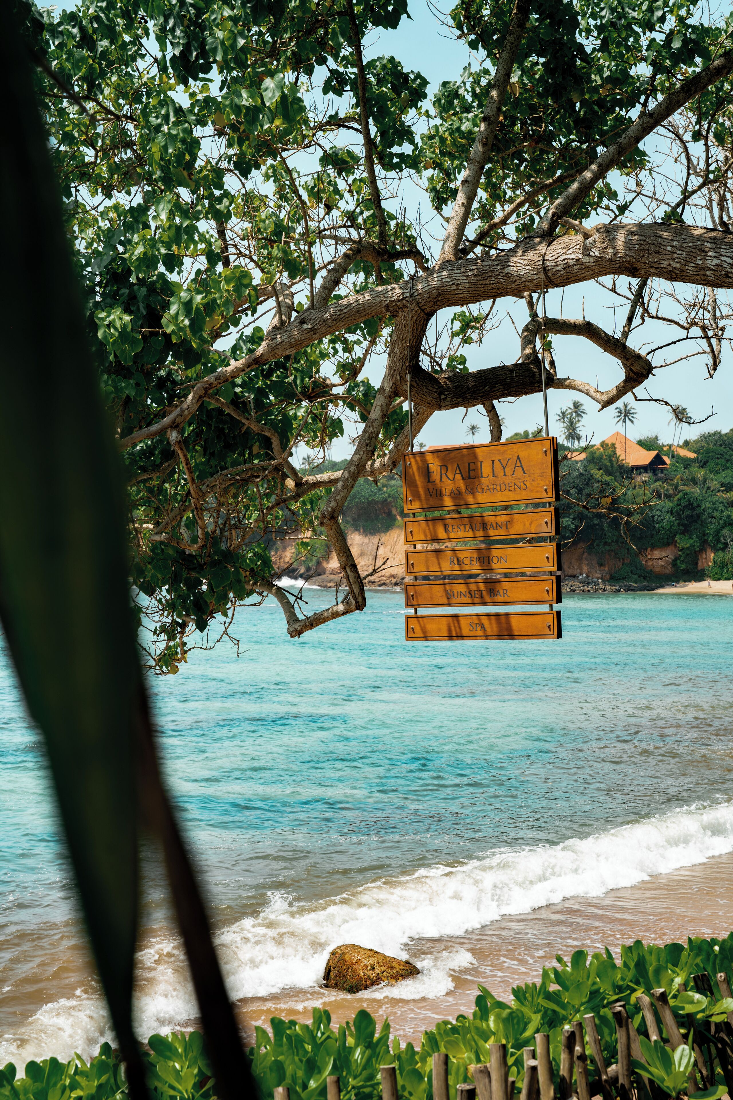 A wooden signboard with 'ERAELIYA VILLAS & GARDENS' and amenities listed, hanging from a tree by the beach.