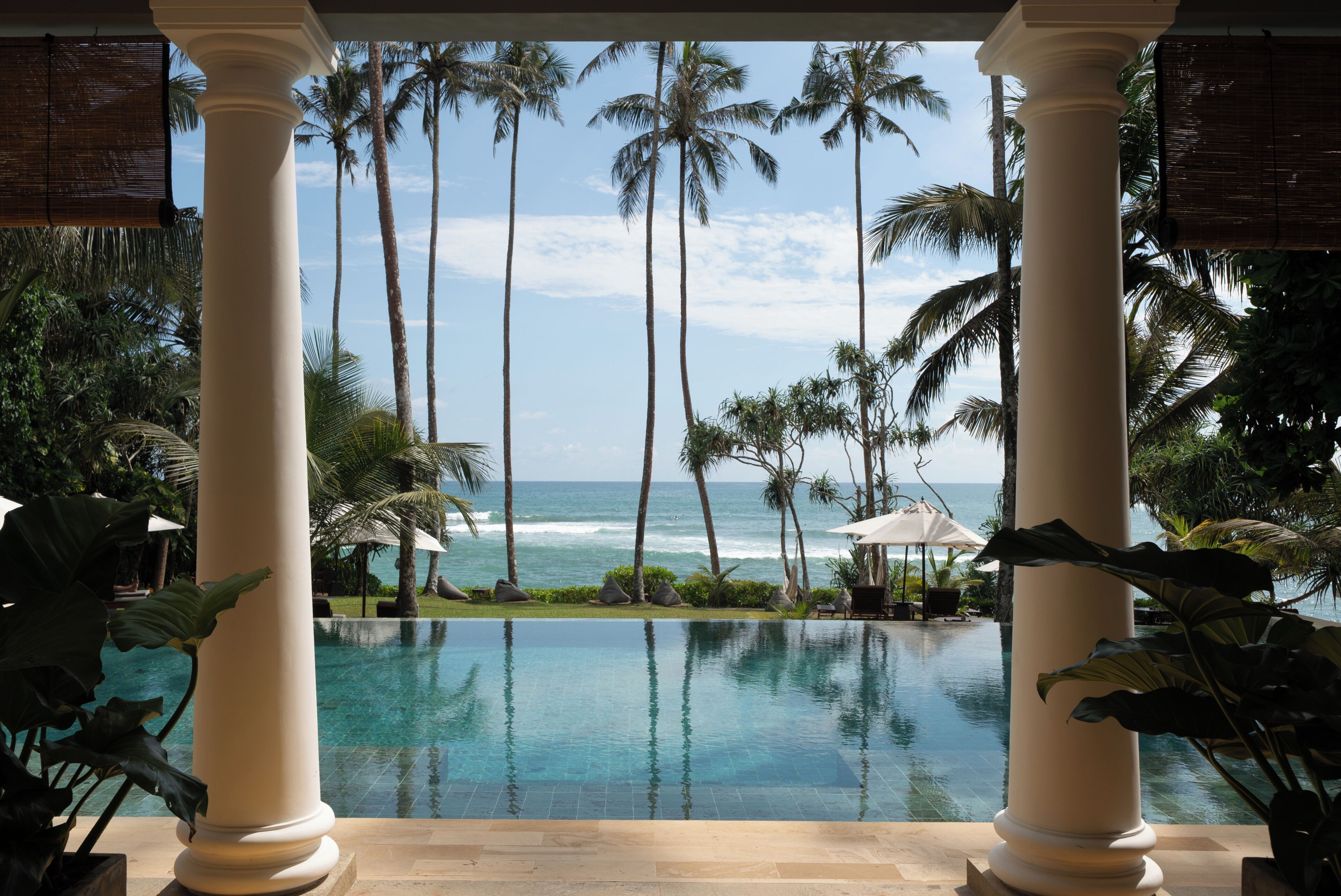 A pool with a view of the ocean, palm trees, and a covered area with columns.