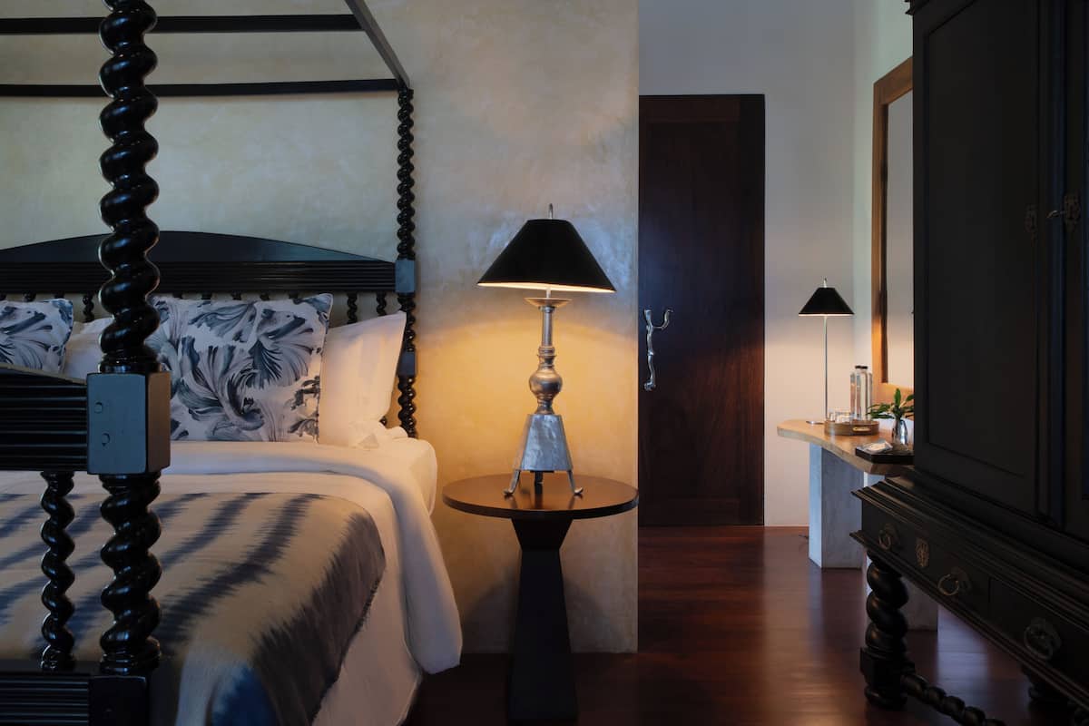 A bedroom with a four-poster bed, a round table with a lamp, a dresser, and a door in the background.