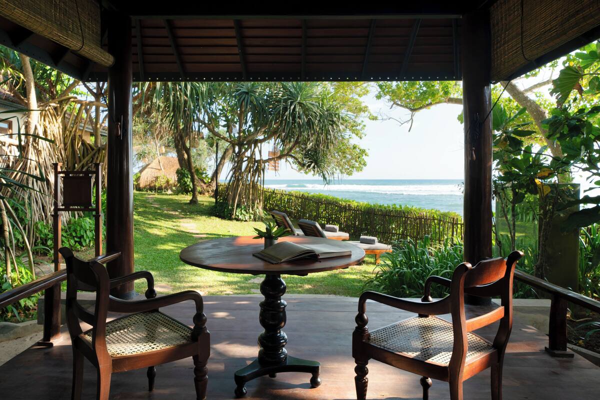 A covered outdoor seating area with a table and chairs, overlooking a lush garden and the ocean.