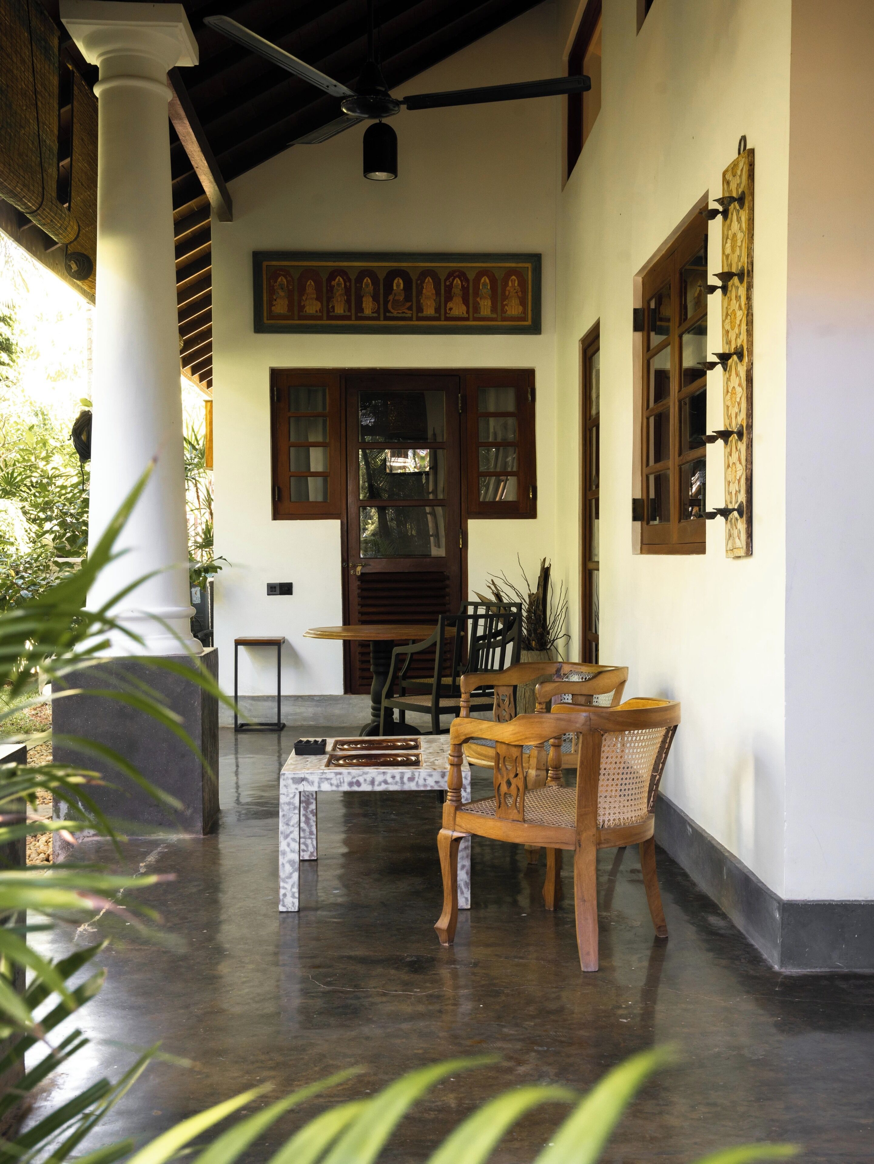 Een overdekt terras met houten stoelen en tafels, een decoratieve muur met religieuze kunst en een groot raam met meerdere ruiten.