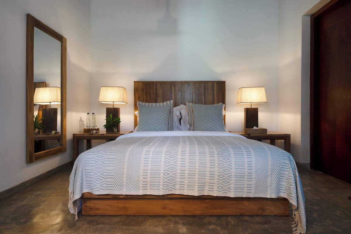 A bedroom with a wooden headboard, two bedside tables with lamps, a large mirror, and a patterned bedspread.