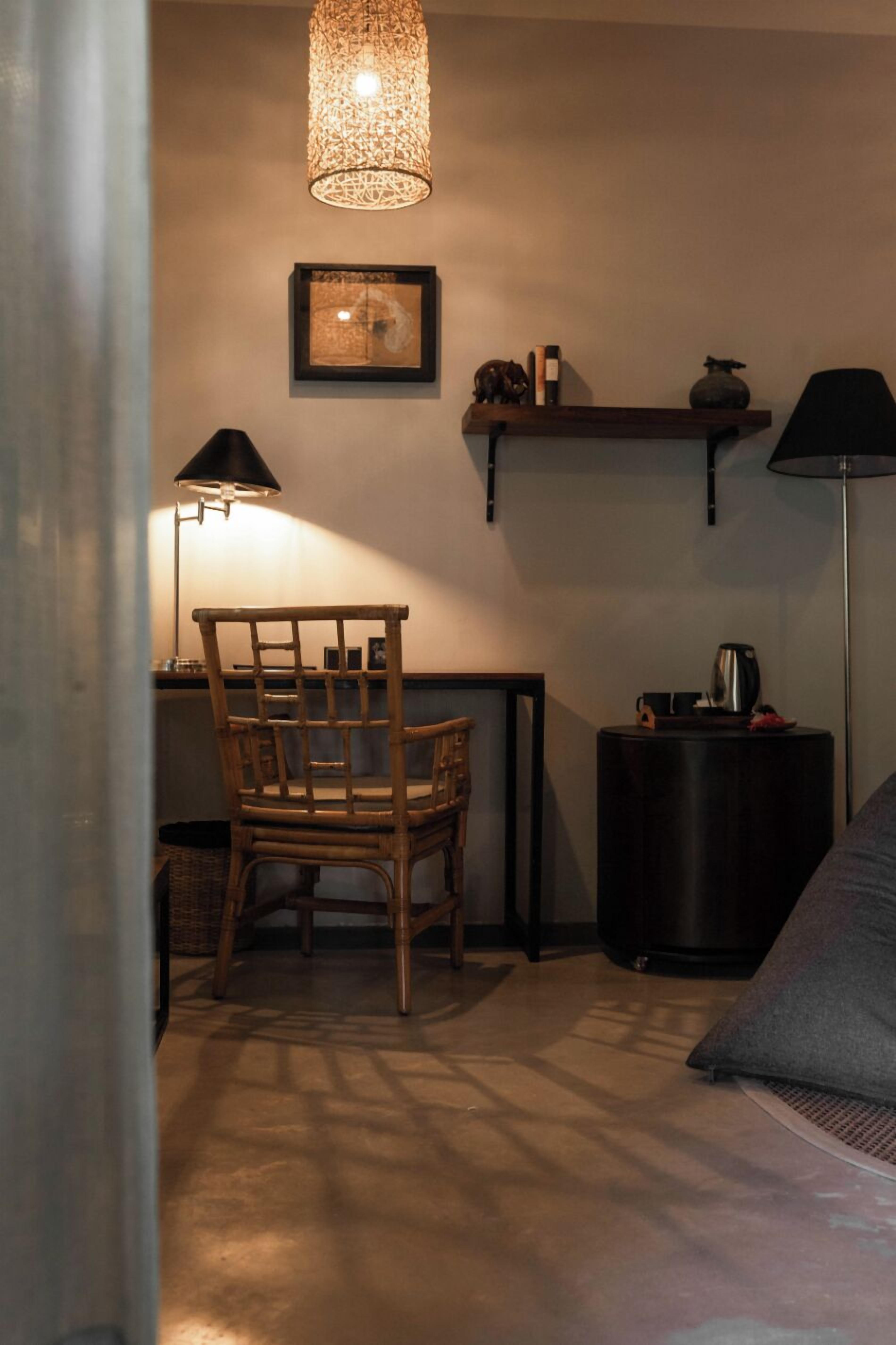 Una habitación con una silla de madera, un escritorio, una lámpara, una estantería con objetos decorativos y una lámpara de pie.