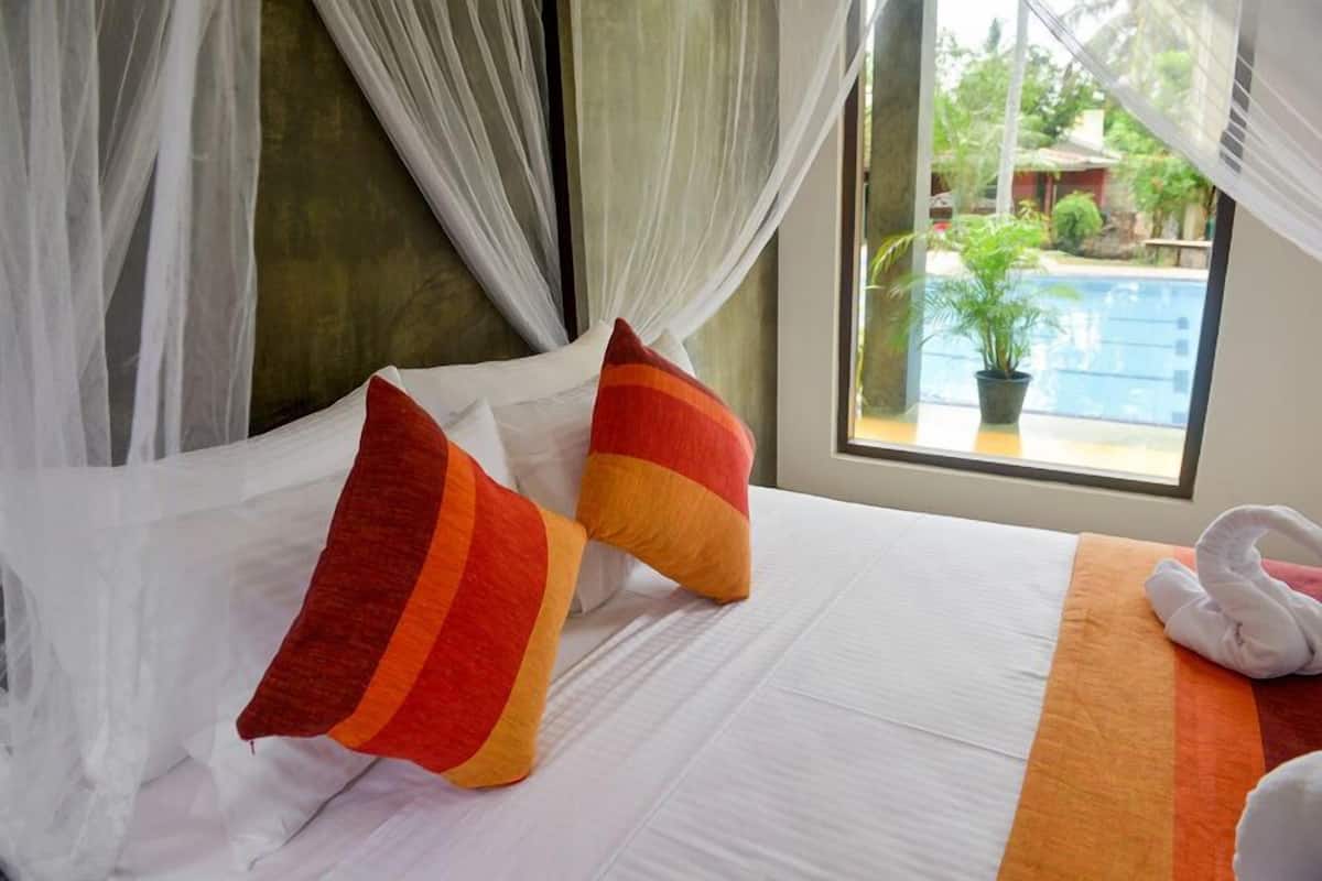 Una cama con sábanas blancas, dos almohadas a rayas y una toalla blanca, con vistas a una piscina y follaje exterior.